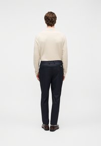 Svetlo bež pulover in temno modre hlače s sproščenim krojem. Vidna pasnica in nežni šivi. Rjavi čevlji zaključujejo videz.