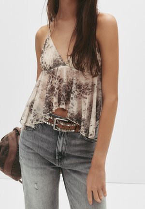 Kobieta ubrana w luźny, bezrękawnik z efektem tie-dye, z brązowym paskiem ozdobionym ćwiekami i wyblakłymi szarymi dżinsami, trzymająca brązowy kapelusz.