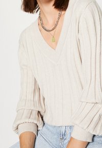 Pull beige clair côtelé avec un col en V, manches bouffantes et texture tricotée. Collier chaîne argentée avec un pendentif en forme d'ourson en gelée vert.