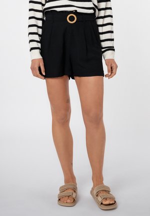 Frau trägt schwarze High-Waist-Shorts mit einem beigen Gürtel mit runder Schnalle, beige geflochtene Sandalen und ein langärmeliges schwarz-weiß gestreiftes Oberteil.