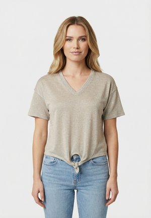 Donna con capelli biondi indossa una maglietta beige a maniche corte con scollo a V annodata e jeans azzurri, in piedi contro uno sfondo chiaro e uniforme.