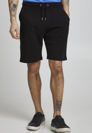 Shorts en coton noirs avec une taille élastique et un cordon de serrage. Équipés de poches latérales et d'un design à longueur de genou. Portés avec des baskets noires.