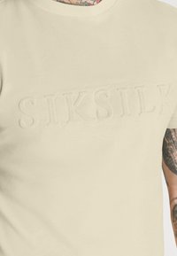 Bežové bavlněné tričko s embosovaným logem "SIKSILK" na přední straně, s kulatým výstřihem a krátkými rukávy, hladká textura.