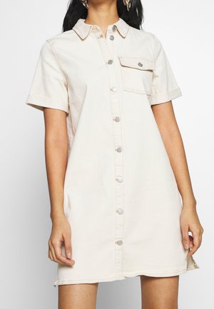 Frau trägt ein beige-farbenes Jeanskleid mit kurzen Ärmeln, Knopfleiste, Kragen und einer Brusttasche.