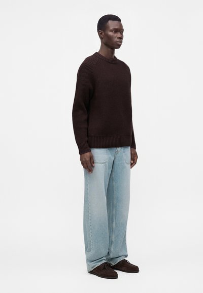Homme debout portant un pull en tricot marron foncé, un jean large bleu clair et des chaussures marron foncé sur un fond clair uni.