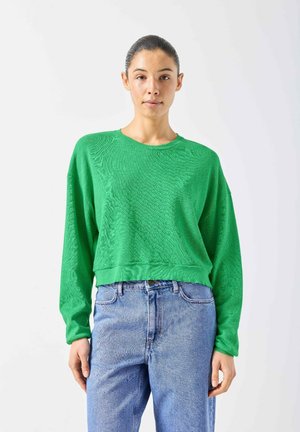 Vrouw met donker haar in een staart, gekleed in een felgroene top met lange mouwen en lichtblauwe jeans met hoge taille, staande tegen een witte achtergrond.