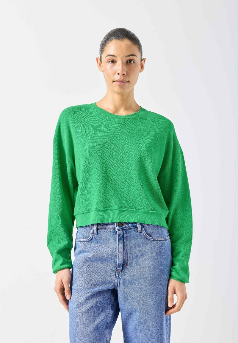 Sieviete ar tumšiem matiem, kas sasieti atpakaļ, valkājot spilgti zaļu garu piedurkņu topu un augsta vidukļa gaiši zilas džinsas, stāvot pret baltu fonu.