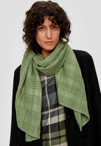 Écharpe verte texturée avec un motif à carreaux, enroulée autour du cou, présentant un matériau doux et un design oversize.