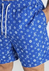 Blå simshorts med vita grafiska mönster, inklusive bokstäver och symboler. Har ett elastiskt midjeband och snörning.