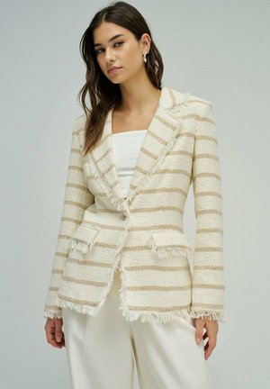 Femme portant un blazer à franges crème et beige à rayures, par-dessus un haut blanc et un pantalon blanc, debout devant un fond clair uni.