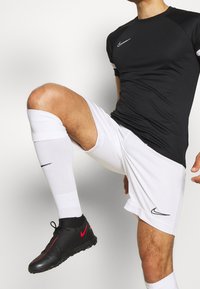 Camiseta deportiva negra ajustada con acentos blancos, combinada con pantalones cortos blancos y calcetas hasta la rodilla. Zapatillas deportivas negras y rojas con diseño texturizado.