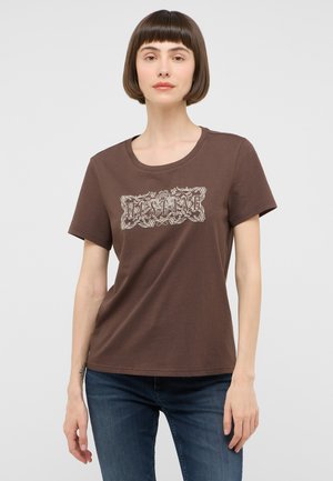 Frau mit kurzen braunen Haaren, die ein braunes T-Shirt mit weißem Blumenmotiv und blaue Jeans trägt, steht vor einem einfachen hellen Hintergrund.