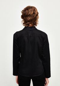 Maglietta nera a maniche lunghe con colletto, caratterizzata da una texture liscia e un orlo curvo, vista da dietro. Design semplice senza decorazioni.