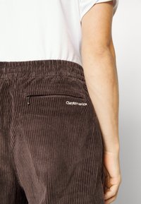 Hnědé kordové kalhoty s vertikálním žebrováním, mají elastický pas a malou kapsu se zipem vzadu. Logo je vyšívané.