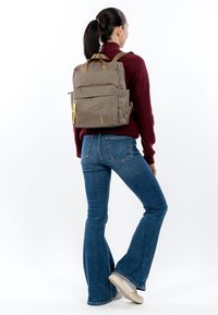 Ruskea kangasreppu, jossa on teksturoitu pinta, kaksi yläkahvaa ja keltainen vetoketjun vedin, ihmisen selässä, joka on pukeutunut rentoihin vaatteisiin.