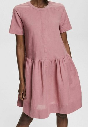 Frau trägt ein locker sitzendes, kurzärmliges pinkes Kleid mit geraffter Taille und rundem Ausschnitt, steht vor einem einfarbigen Hintergrund.