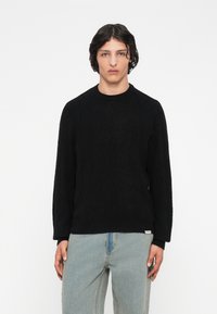 Mladý muž v čiernom štruktúrovanom pletenom svetre a svetlomodrých džínsoch, stojaci na pozadí bielej steny.