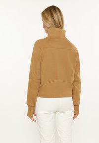 Felpa marrone con colletto alto, polsini a costine e design cropped. Texture liscia con dettagli di cucitura. Indossata sopra pantaloni bianchi.