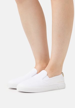 Baskets en toile blanches à enfiler avec une semelle texturée, mettant en avant un accent doré au talon et un branding subtil sur le côté.