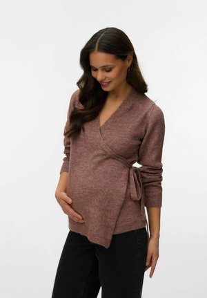 Rase naine pikkade tumedate juustega, kellel on seljas lilla mähissviiter ja mustad püksid, hoides õrnalt oma kõhtu ja naeratades alla.