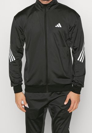 Mężczyzna ubrany w czarny dres marki Adidas z białym logo na klatce piersiowej oraz trzema białymi paskami na rękawach i spodniach, stoi z rękami swobodnie opuszczonymi.