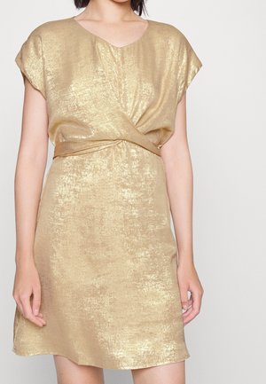 Kobieta ubrana w błyszczącą złotą sukienkę z krótkim rękawem i skręconym detalem w talii, stojąca na tle jednolitego jasnego tła.