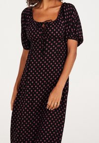 Svart klänning med korta puffärmar, med en rundad halsringning och dragsko. Tyget har ett rosa diamantmönster genom hela.