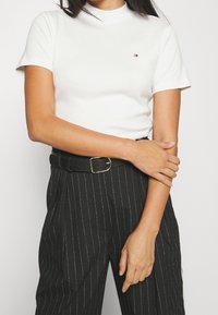 Bílá bavlněná tričko s krátkými rukávy, vysokým výstřihem a malým logem. Kombinováno s černými kalhotami s proužky a černým opaskem.