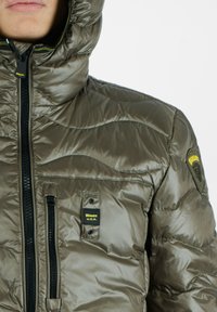 Gepolsterte Kapuzenjacke in Olivgrün, mit einem vorderen Reißverschluss, Seitentaschen und strukturiertem Stoff mit gesteppten Wellenmustern. Schwarze Hardware-Akzente.