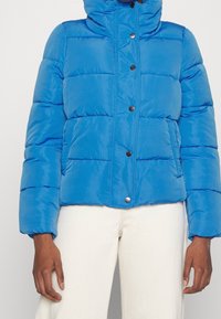 Blå puffjacka med hög krage, framsnapplikation och horisontellt quiltat mönster; har en slät textur och elastiska ärmmuddar.