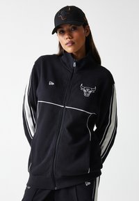 Svart zip-up jakke med hvite striper, med Chicago Bulls-logo på brystet. Kombinert med en svart caps med brodert logo.