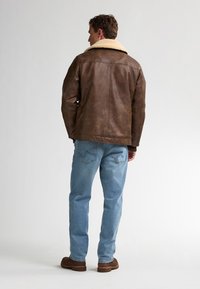 Hombre vestido con una chaqueta marrón de cuello de borrego, jeans azul claro y botas marrones, de pie y mirando hacia atrás contra un fondo liso.