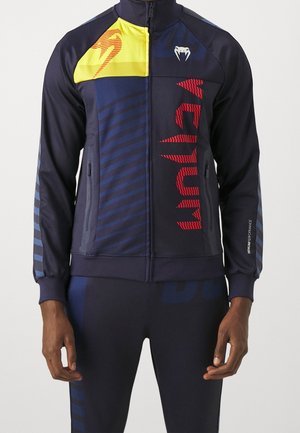 Mężczyzna ubrany w granatową sportową kurtkę z czerwonym napisem "Venum", żółty panel na ramieniu z logo oraz dopasowane granatowe spodnie z paskami.