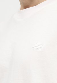 Κοντινό πλάνο ενός ατόμου που φοράει λευκό μπλουζάκι με μικρό, ανάγλυφο λογότυπο New Balance στο αριστερό στήθος.