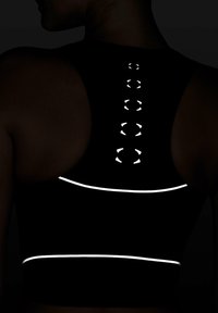 Sort sports-bh med refleksdetaljer langs ryggen og bunden. Har et racerback-design med en række cirkulære refleksdetaljer.