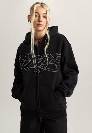 Młoda osoba o długich blond włosach w czarnej bluzie z kapturem z białym abstrakcyjnym wzorem, stojąca z rękami w kieszeniach na jednolitym tle.