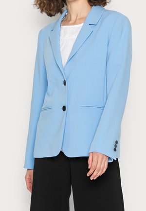 Femme portant un blazer bleu clair cintré avec des boutons noirs, un top blanc texturé et un pantalon noir, debout devant un fond uni.