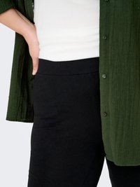 Camicia verde a bottoni con un tessuto texture, indossata sopra una maglia bianca, abbinata a pantaloni neri aderenti. Focus sul look a strati dell'abbigliamento.