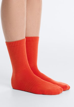 Beine mit leuchtend orangefarbenen, gerippten Crew-Socken, die auf einer hellgrauen Oberfläche mit schlichtem weißen Hintergrund stehen.
