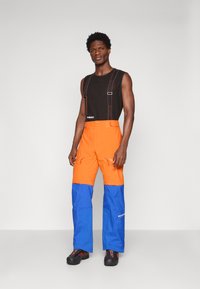 Άνδρας που φοράει μαύρο αμάνικο μπλουζάκι με πορτοκαλί και μπλε παντελόνια εξωτερικού χώρου με τιράντες, σε φόντο λευκό.