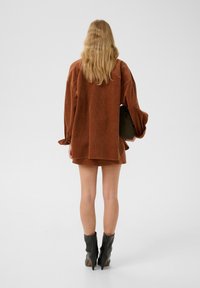 Camicia oversized in corduroy marrone con maniche lunghe, abbinata a una mini gonna coordinata. Stivaletti neri alla caviglia con punta allungata, mentre si tiene una borsa verde.