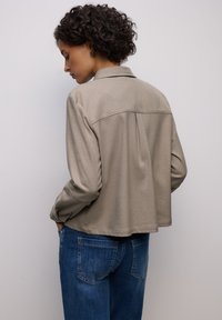 Beige Jacke mit Hemdkragen und langen Ärmeln, mit lockerem Schnitt und strukturiertem Stoff, kombiniert mit blauen Jeans.