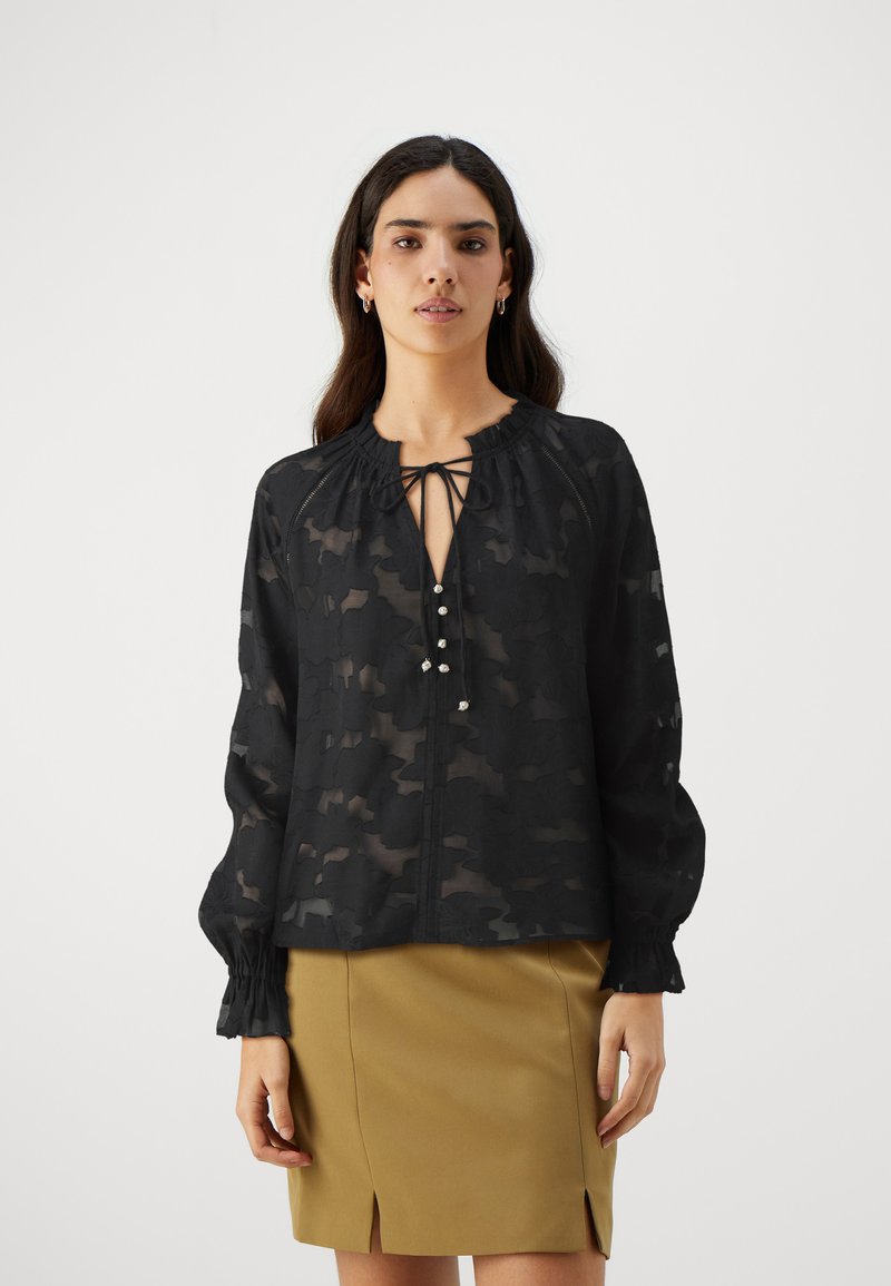 Blusa nera trasparente con motivo floreale, scollatura a V profonda, dettaglio con fiocco e bottoni decorativi. Presenta polsini arricciati e vestibilità ampia.
