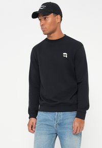 Svart sweatshirt med lange ermer, ribbestrikkede mansjetter og hem. Har en liten hvit grafikk på brystet. Kombinert med lyseblå jeans.