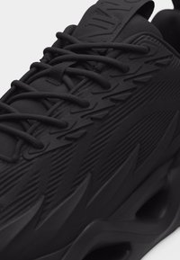 Κοντινό πλάνο μαύρου αθλητικού παπουτσιού με υφή στο πάνω μέρος, περίπλοκο σχέδιο κορδονιών και παχύ γλυπτό πέλμα με γωνιώδεις αυλακώσεις και ανοίγματα.