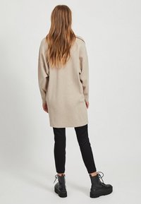Femme aux longs cheveux portant une tunique beige à manches longues, un pantalon noir slim et des bottines noires à lacets, debout devant un fond uni.