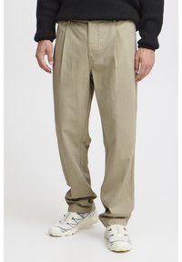 Pantalon beige ample avec des plis à l'avant, fabriqué en tissu doux. Les coutures latérales présentent des surpiqûres contrastantes. Porté avec des baskets blanches.