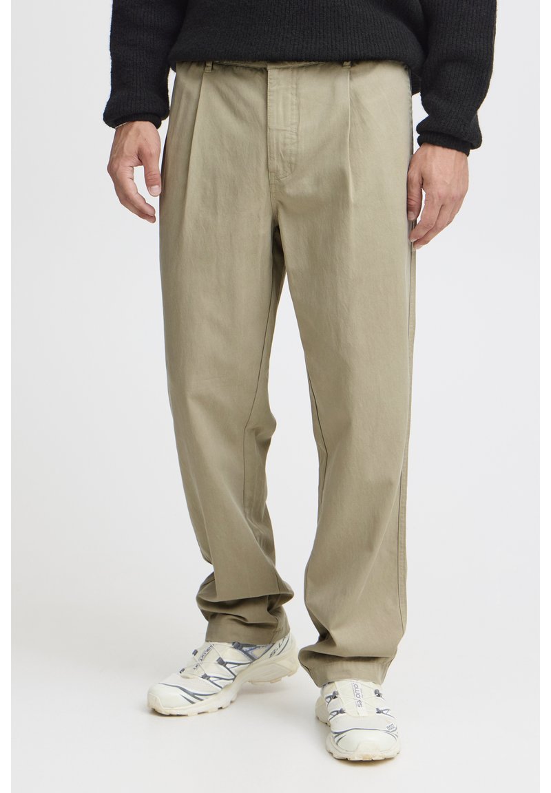 Pantalon beige ample avec des plis à l'avant, fabriqué en tissu doux. Les coutures latérales présentent des surpiqûres contrastantes. Porté avec des baskets blanches.