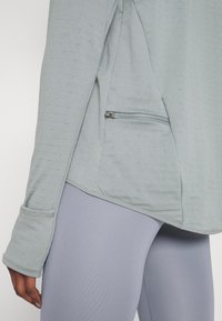 Lichtgrijze top met lange mouwen en ribbeltextuur, voorzien van een zijzak met rits. Gecombineerd met lavendel leggings, die de gestroomlijnde vorm benadrukken.