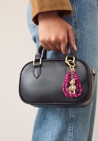 Zwarte leren handtas met afgeronde vorm, gouden hardware en een roze gevlochten hanger met een klein metalen beertje. Hand houdt de tas vast.